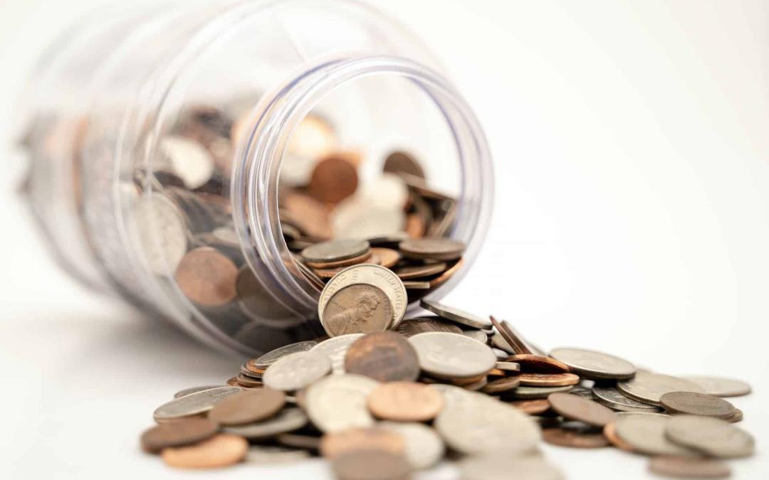 A clear plastic jar tipped over, spilling a variety of coins, including nickels, dimes, and pennies onto a white surface. The coins correlate with the 2021 HTF Income Limits, now scattered around the overturned jar.