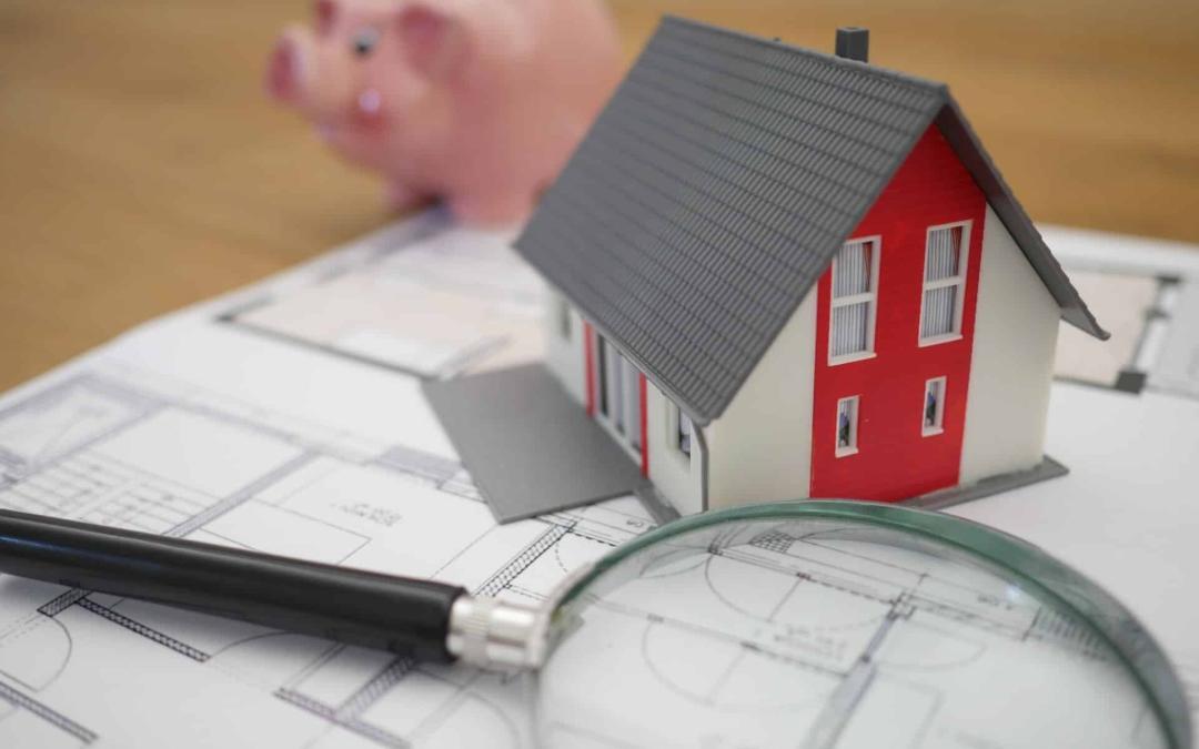 A miniature house model with red and white walls sits on architectural blueprints labeled