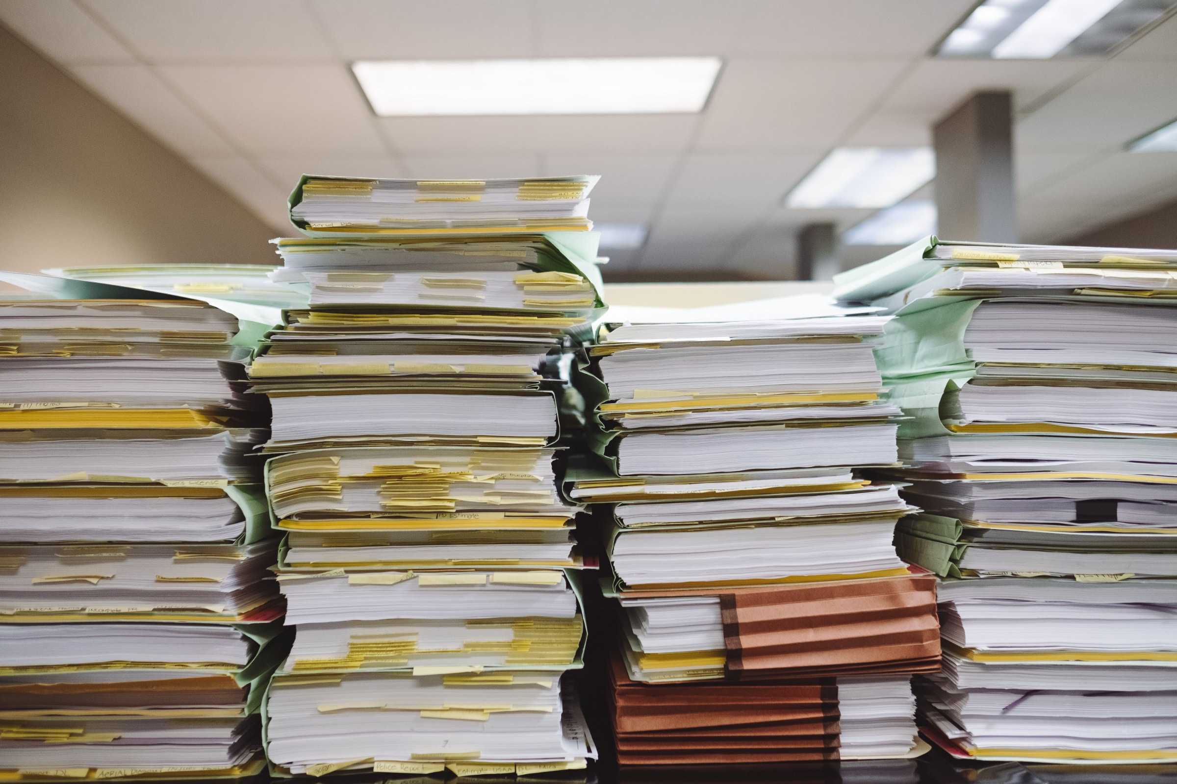 Stacks of organized documents and folders on a desk under fluorescent office lighting. The papers are neatly arranged in various colored folders, indicating a large volume of paperwork. The background shows a typical office ceiling with light fixtures.